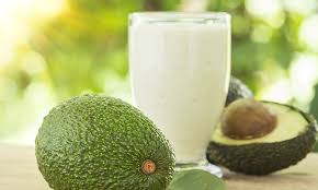 Image of a cup of Avocado Lassi in a tumbler along with a whole and cut avocado fruit displayed